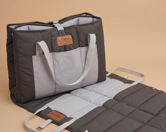 Puede incluir: Un bolso transportín acolchado para mascotas en gris oscuro y gris claro con una alfombrilla desplegable. El bolso cuenta con dos asas gris claro, un cierre de hebilla superior y dos parches de cuero sintético color canela con Charlie y LUNNA.