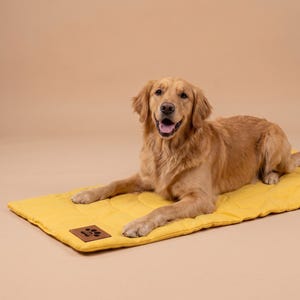 May include: A golden retriever dog lying on a rectangular yellow quilted pet mat. The mat features a brown rectangular leather-like patch in the corner with a black paw print graphic embossed on the surface, providing a soft resting area.