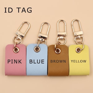 May include: Four rectangular ID tags with gold-tone metal lobster clasps and rings. The tags are pink, blue, brown, and yellow, each featuring its respective color name printed in black text. The words ID TAG appear in the upper left corner.