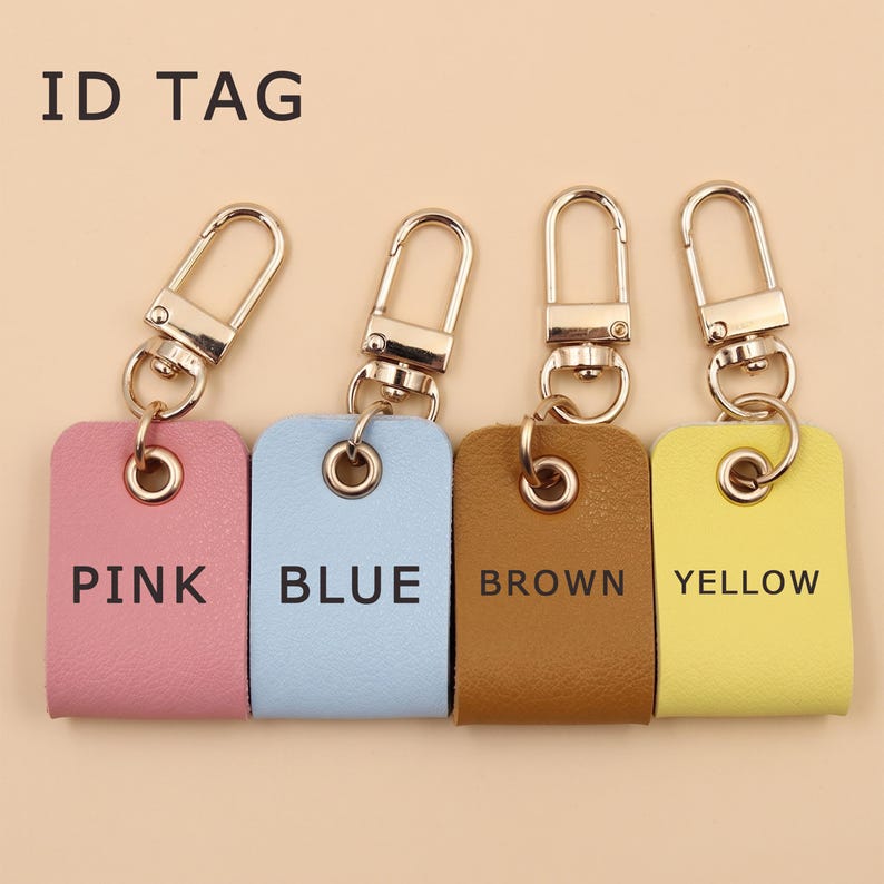 May include: Four rectangular ID tags with gold-tone metal lobster clasps and rings. The tags are pink, blue, brown, and yellow, each featuring its respective color name printed in black text. The words ID TAG appear in the upper left corner.