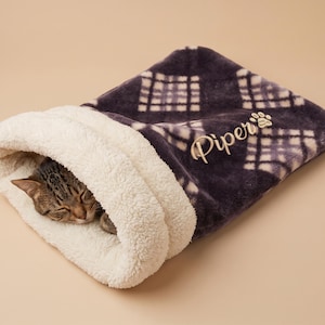 Puede incluir: Un saco de dormir de felpa para mascotas con un patrón de cuadros morado oscuro y blanco en el exterior y un forro de forro polar sherpa color crema. El nombre Piper está bordado en cursiva con un pequeño icono de huella en la parte superior.