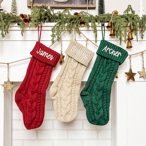 May include: Three cable-knit Christmas stockings in red, cream, and green hang from a garland. Each stocking has a folded cuff and a name embroidered in white: "James", "Brooke", and "Archer". The stockings are suspended by red and green loops.