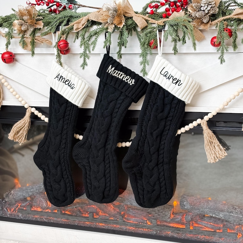 May include: Three black cable knit Christmas stockings with white cuffs hang above a fireplace. Each stocking is personalized with a name embroidered in white: Amelia, Matthew, and Lauren. Festive greenery and red berries decorate the mantel.
