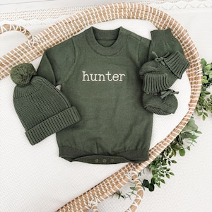Peut inclure: Une barboteuse &agrave; manches longues en tricot vert olive avec le mot hunter brod&eacute; en blanc sur la poitrine, accompagn&eacute;e d'un bonnet assorti avec pompon et d'une paire de chaussons en tricot vert olive avec des rubans aux chevilles.
