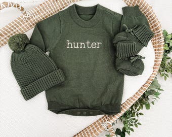 May include: An olive green baby outfit set, including a knit romper with the name "hunter" embroidered in white, a matching beanie with a pom-pom, and booties. The set is displayed in a woven basket with greenery.