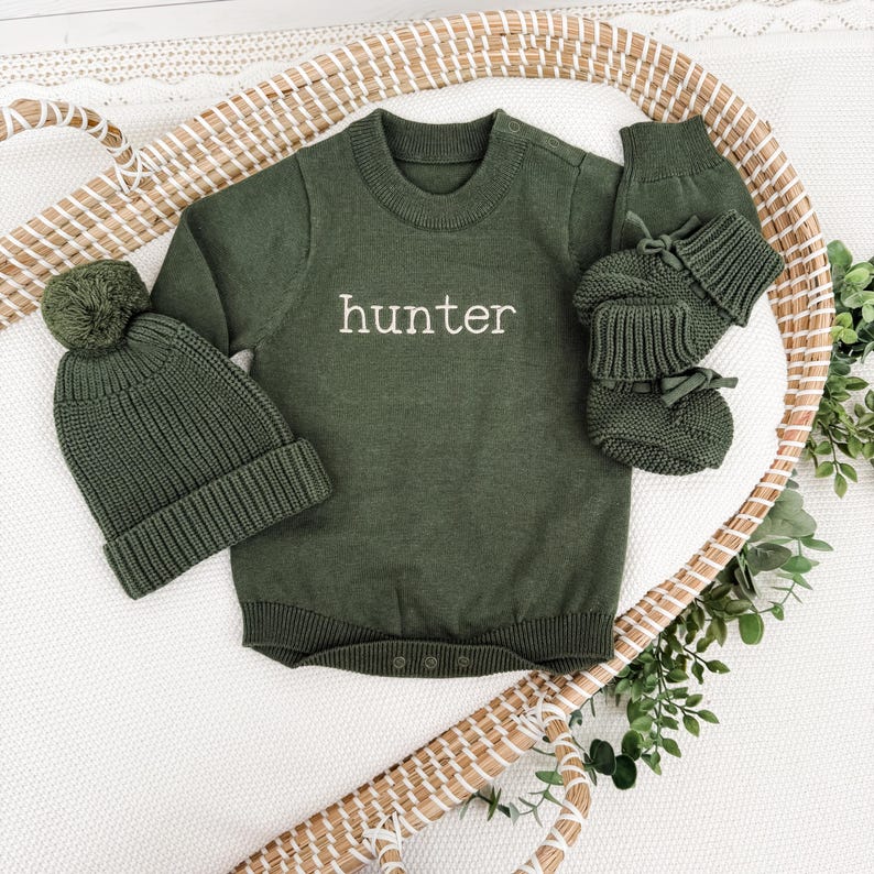 Peut inclure: Une barboteuse &agrave; manches longues en tricot vert olive avec le mot hunter brod&eacute; en blanc sur la poitrine, accompagn&eacute;e d'un bonnet assorti avec pompon et d'une paire de chaussons en tricot vert olive avec des rubans aux chevilles.