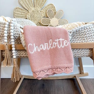 May include: A pink baby blanket with the name "Charlotte" embroidered in white script. The blanket has a scalloped edge and is draped over a woven bassinet. Decorative wooden flowers and a beaded garland are in the background.