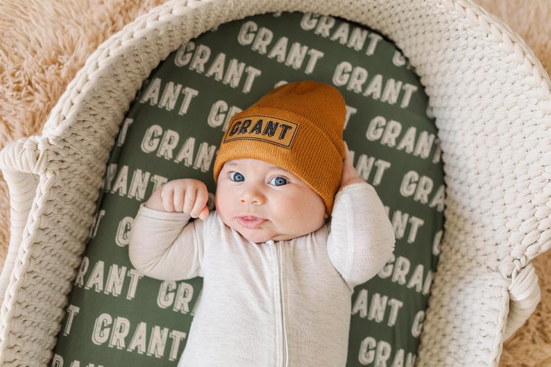 May include: A baby wearing a brown knit beanie with the word "GRANT" embroidered on it. The baby is lying in a white wicker basket lined with a green fabric with the word "GRANT" repeated in white.
