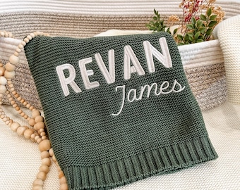 May include: A dark olive green knitted blanket featuring the name REVAN James embroidered in white capital and script lettering. The blanket has a ribbed hem and is accompanied by a strand of light-colored wooden beads.
