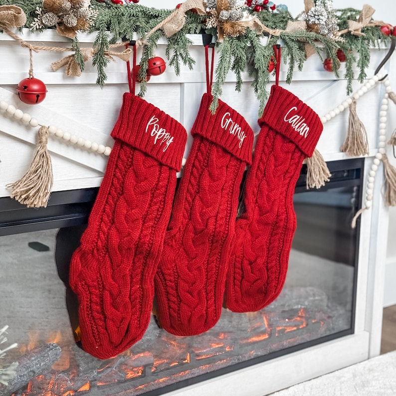 May include: Three red cable-knit Christmas stockings hang from a white mantel. Each stocking is personalized with a name in white embroidery: Poppy, Grimy, and Callan. The mantel is decorated with greenery and red bells.