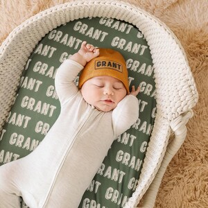 May include: A baby wearing a white sleeper and a brown knit hat with the word "GRANT" embroidered on it. The baby is sleeping in a white wicker bassinet with a green and white patterned sheet that says "GRANT" repeatedly.