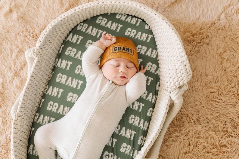 May include: A baby wearing a white sleeper and a brown knit hat with the word "GRANT" embroidered on it. The baby is sleeping in a white wicker bassinet with a green and white patterned sheet that says "GRANT" repeatedly.