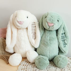 Puede incluir: Dos conejos de peluche, uno crema y otro verde salvia, se muestran juntos. El conejo crema tiene el nombre "Aaliyah" bordado en su oreja, mientras que el conejo verde tiene "Ellie" bordado en su oreja. Ambos conejos tienen narices rosas y pelaje suave y esponjoso.