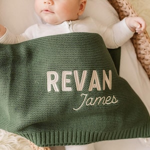 May include: A green knitted baby blanket with the name "REVAN James" embroidered in white. The blanket is draped over a baby in a wicker bassinet, creating a cozy and personalized nursery item.