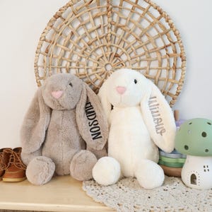 May include: Two plush bunny rabbits, one gray and one white, with long floppy ears and pink noses. The gray bunny has the name "HUDSON" embroidered on its ear. The white bunny has the name "Julietas" embroidered on its ear. They are sitting on a wooden surface.