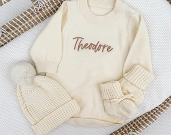 May include: A cream-colored long-sleeved knit sweater with the name Theodore embroidered in brown script on the chest, accompanied by a matching ribbed knit beanie with a pom-pom and a pair of soft knit baby booties with ribbon ties.