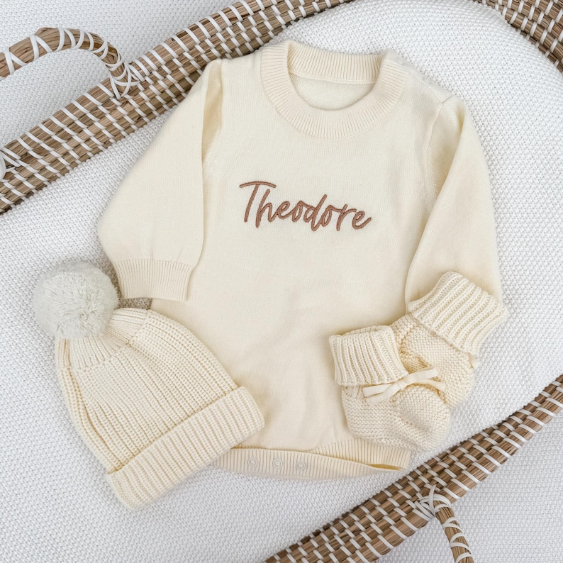 Peut inclure: Un pull &agrave; manches longues en tricot couleur cr&egrave;me avec le pr&eacute;nom Theodore brod&eacute; en lettres cursives marron sur la poitrine, accompagn&eacute; d'un bonnet en tricot c&ocirc;tel&eacute; assorti avec pompon et d'une paire de chaussons en tricot avec un petit n&oelig;ud.