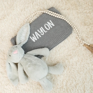 May include: A gray plush bunny toy with long ears and a pink nose rests next to a gray knit blanket. The blanket has the name "WAYLON" in white lettering. A beaded garland with a tassel is also present.