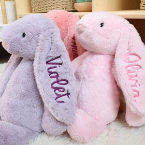 May include: Three plush bunny rabbits in shades of lavender, pink, and rose. The lavender bunny has "Violet" embroidered on its ear in purple thread. The pink bunny has "Olivia" embroidered on its ear in pink thread. The bunnies are sitting on a white surface.