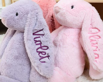 May include: Three plush bunny rabbits in shades of lavender, pink, and rose. The lavender bunny has "Violet" embroidered on its ear in purple thread. The pink bunny has "Olivia" embroidered on its ear in pink thread. The bunnies are sitting on a white surface.