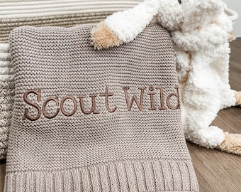 May include: A light brown knitted blanket featuring the embroidered text Scout Wild in a dark brown script font, paired with a white plush stuffed animal with long ears and tan accents, all resting inside a woven rope storage basket.