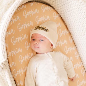 May include: A baby wearing a cream-colored beanie with a gold "Sophia" name tag, lying on a mustard yellow and white patterned fabric. The fabric has the name "Sophia" written repeatedly in white.
