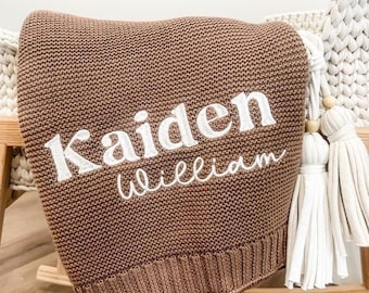 May include: A brown knitted baby blanket with the name "Kaiden William" embroidered in white. The blanket is draped over a wooden rocking cradle. White tassels hang from the side of the cradle.