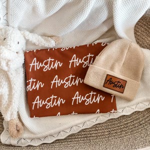 May include: A beige beanie with a brown leather patch that reads "Austin" sits next to a rust-colored pillow with the name "Austin" printed in white. A white blanket and a stuffed animal are also in the image.