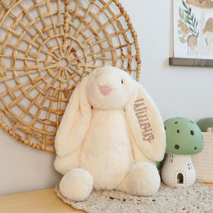 May include: A large, cream-colored plush bunny with long, floppy ears and the name "Willis" embroidered on one ear. The bunny has a pink nose and is sitting on a beige crocheted mat. A green mushroom-shaped night light is next to the bunny.