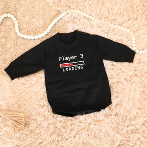 May include: A black long-sleeved baby bodysuit featuring the white text Player 3 and a loading bar graphic with red segments. The garment has a round neckline and snap closures at the shoulder and bottom for easy dressing and diaper changes.
