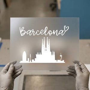 May include: A clear, rectangular panel with a frosted finish, featuring a white silhouette of the Barcelona skyline. The word "Barcelona" is written in cursive script above the skyline, with a heart symbol. The panel is held by gloved hands.