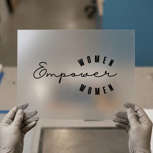 May include: A frosted clear acrylic sign with the words "Empower Women" in black script and block letters. The sign is held by gloved hands against a neutral background.