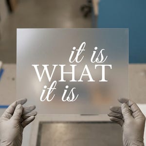 May include: A frosted glass panel with the phrase "it is WHAT it is" in white lettering. The panel is held by gloved hands against a blurred background, suggesting a workshop or studio setting.