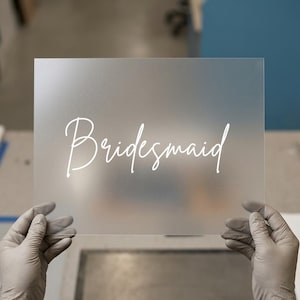May include: A frosted acrylic sign with the word "Bridesmaid" in elegant white script. The sign is rectangular and held up by gloved hands, with a blurred background.