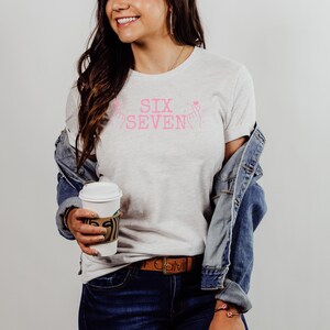 Model wearing a Bella+Canvas 3001 unisex t-shirt in Ash. The front features soft pink distressed text reading “SIX SEVEN,” flanked by illustrated finger heart hand gestures with small heart accents. The shirt is styled casually with a denim jacket, jeans, and a coffee cup, shown in a clean studio setting.