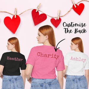 Back view mockup showing three Bella+Canvas 3001 unisex t-shirts in Black Heather, Heather Charity Pink, and Ash. Each shirt features a custom name printed across the upper back in coordinating pink lettering. The image includes decorative red heart cutouts clipped on string above the shirts and text that reads “Customize the Back,” highlighting the personalized name option.
