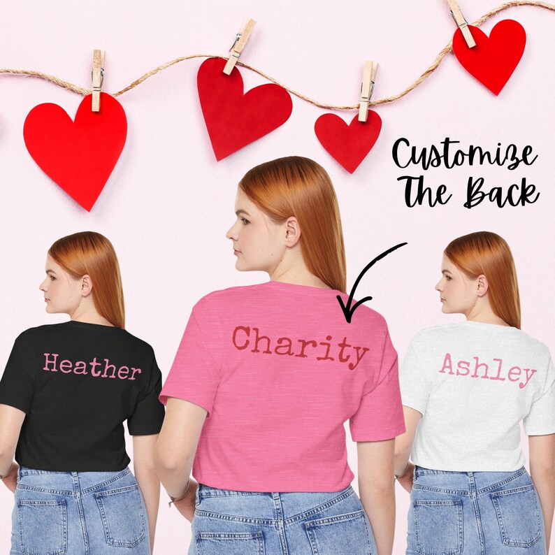 Back view mockup showing three Bella+Canvas 3001 unisex t-shirts in Black Heather, Heather Charity Pink, and Ash. Each shirt features a custom name printed across the upper back in coordinating pink lettering. The image includes decorative red heart cutouts clipped on string above the shirts and text that reads “Customize the Back,” highlighting the personalized name option.