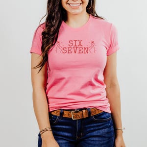 Model wearing a Bella+Canvas 3001 unisex t-shirt in Heather Charity Pink. The front features red distressed text reading “SIX SEVEN,” flanked by illustrated finger heart hand gestures with small heart accents. The shirt is styled with jeans and simple jewelry in a clean studio setting.