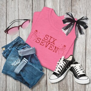 Flat lay of a Bella+Canvas 3001 t-shirt in Heather Charity Pink featuring red “SIX SEVEN” text with illustrated finger heart hand gestures and small heart accents. The shirt is styled with folded denim shorts, black low-top sneakers, pink-tinted sunglasses, and a sheer black bow on a light wood background, creating a trendy pop-inspired outfit layout.