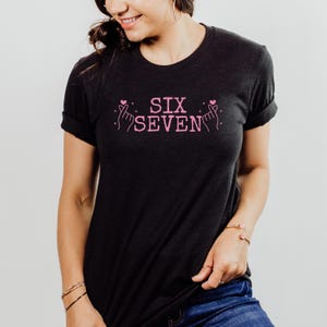 Model wearing a Bella+Canvas 3001 unisex t-shirt in Black Heather. The front features pink distressed text reading “SIX SEVEN,” flanked by illustrated finger heart hand gestures with small heart accents. The shirt is styled casually with rolled sleeves, jeans, and simple jewelry, shown in a clean studio setting.