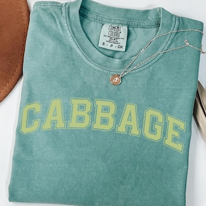 May include: A seafoam green t-shirt with the word "CABBAGE" printed in a light green, block letter font. The shirt has a crew neck and short sleeves. A silver necklace with a small pendant is draped across the shirt.