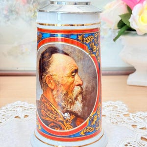 Peut inclure: Une chope à bière en céramique blanche avec un couvercle et une poignée en argent. La chope présente un portrait d'un homme barbu, encadré d'éléments décoratifs rouges, bleus et dorés. La chope a un bord doré et est probablement un objet de collection.