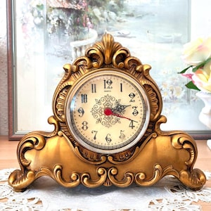 May include: A gold-colored mantel clock with an ornate, curved frame and a cream-colored face. The clock features black Arabic numerals, decorative central filigree, and black hour and minute hands with a thin red second hand. The word Quartz is printed below the number 12.