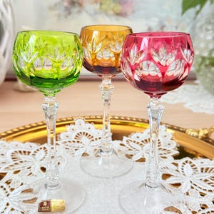 Puede incluir: Tres copas de vino de cristal de colores. Las copas tienen un tallo y una base transparentes, con un cuenco redondo en verde, ámbar y rojo. Cada cuenco presenta un patrón en forma de hoja. Las copas se exhiben en una bandeja dorada con un tapete de encaje blanco.
