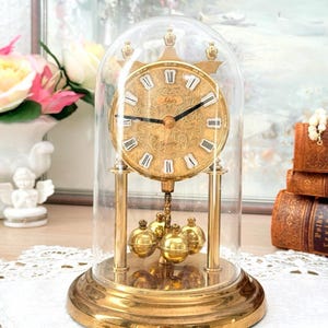 Può includere: Un orologio al quarzo dorato con una cupola di vetro. Il quadrante dell'orologio presenta numeri romani e il marchio "Schatz". L'orologio ha un pendolo con cinque sfere dorate e poggia su una base rotonda. È un oggetto d'antiquariato.