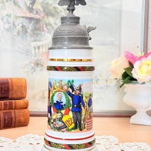 Può includere: Un boccale di birra decorativo in ceramica con un'illustrazione dettagliata di soldati in uniforme blu. Il boccale presenta un coperchio in metallo con un'aquila e fasce decorative rosse, nere e oro. La base ha testo in tedesco.