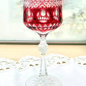 May include: A ruby red crystal wine glass with a detailed cut pattern. The bowl is a deep red, transitioning to clear at the stem. The stem has a decorative ball, and the base is clear with a starburst design.