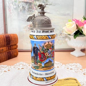 May include: A vintage German beer stein with a detailed metal lid featuring an eagle. The ceramic body is decorated with a military figure, coat of arms, and architectural scenes. The stein has text in German and is trimmed with gold and blue accents.