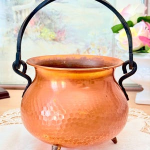 Peut inclure: Un chaudron en cuivre martelé avec un corps arrondi, trois petits pieds et une anse pivotante en métal sombre fixée à deux boucles latérales. Le récipient présente un bord supérieur large et ouvert ainsi qu'une surface texturée et brillante.
