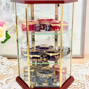 May include: A glass display cabinet with a dark wood base and trim, showcasing various decorative porcelain boxes and thimbles. The boxes are in shades of red, blue, and black, adorned with gold floral designs. The cabinet has multiple glass shelves.